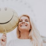Cheerful woman holding a smiley balloon outdoors on a sunny day, exuding happiness and positivity.